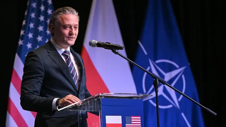 Szef BBN Bartosz Grodecki przemawia do mikrofonu. W tle flaga USA, Polski i NATO
