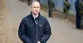 Cezary Tomczyk w kurtce przemawia do mikrofonu. W tle płot graniczny i żołnierze