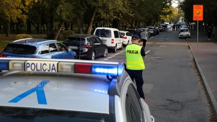 Funkcjonariusz policji, na pierwszym planie radiowóz w tle przejście dla pieszych i zaparkowane samochody