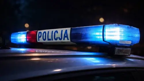 Radiowóz policji z włączonymi niebieskimi i czerwonymi światłami na dachu nocą, widoczny napis „POLICJA” na belce świetlnej.