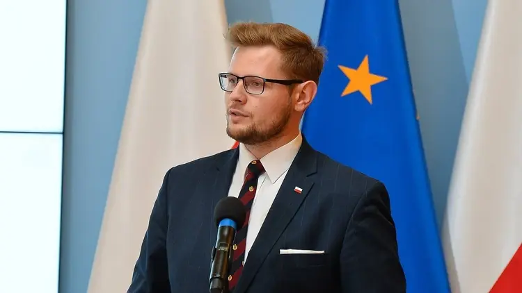 Michał Woś przemawia do mikrofonu na konferencji prasowej. W tle flagi Polski i UE