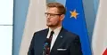 Michał Woś przemawia do mikrofonu na konferencji prasowej. W tle flagi Polski i UE