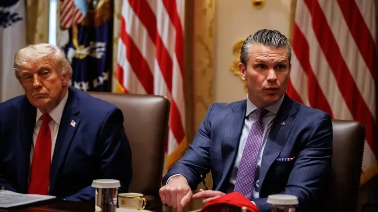 Donald Trump siedzi obok Pete Hegseth, który wskazuje ręką na stół i coś mówi. W tle widoczne są amerykańskie flagi.