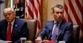 Donald Trump siedzi obok Pete Hegseth, który wskazuje ręką na stół i coś mówi. W tle widoczne są amerykańskie flagi.