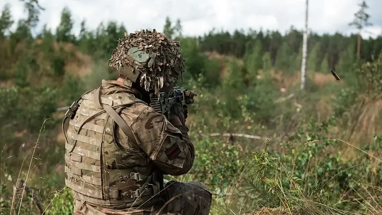 Żołnierz NATO w kamuflażu prowadzi ogień podczas ćwiczeń wojskowych w terenie leśnym