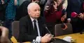 Jarosław Kaczyński siedzi przy biurku i mikrofonie otoczony dziennikarzami