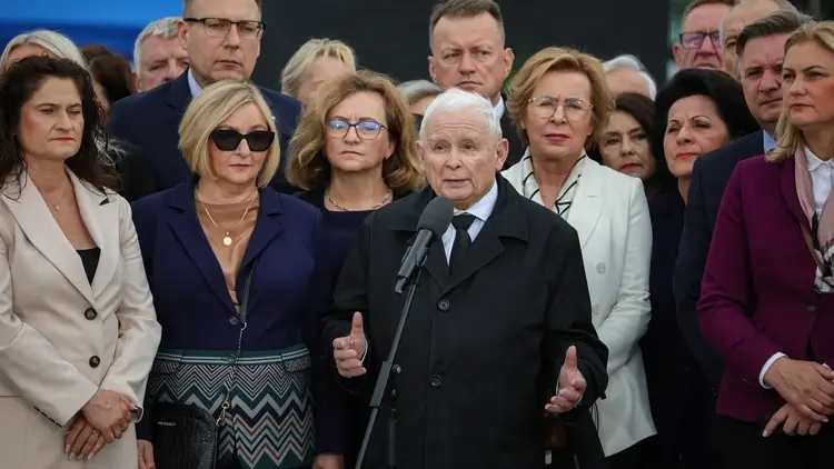 Jarosław Kaczyński przemawia do mikrofonu i gestykuluje. Wokół stoją ludzie
