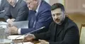 Wołodymyr Zełenski siedzi przy biurku przemawia do mikrofonu. W tle mężczyźni w garniturach