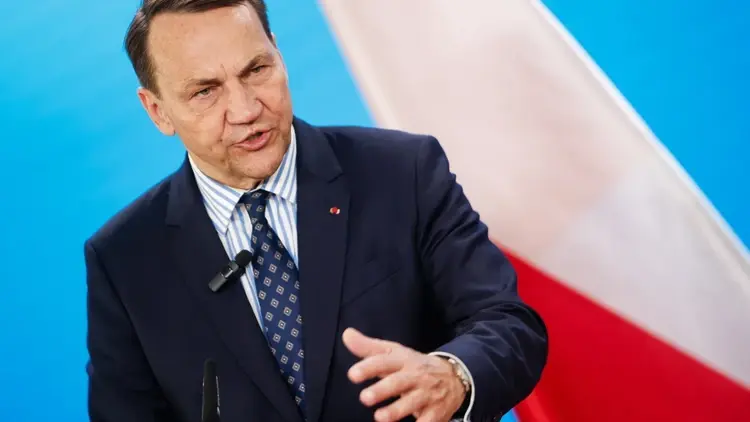 Radosław Sikorski przemawia i gestykuluje. W tle polska flaga