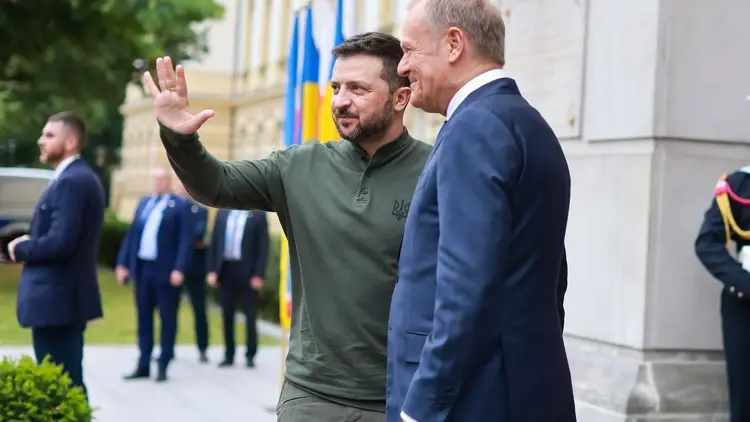 Wołodymyr Zełenski macha ręką, Donald Tusk uśmiecha się. W tle mężczyźni w garniturach