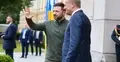 Wołodymyr Zełenski macha ręką, Donald Tusk uśmiecha się. W tle mężczyźni w garniturach