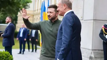Wołodymyr Zełenski macha ręką, Donald Tusk uśmiecha się. W tle mężczyźni w garniturach
