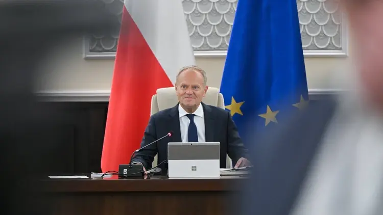 Donald Tusk siedzi przy biurku. W tle flaga Polski i UE