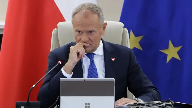 Donald Tusk z ręką przy ustach siedzi w białym fotelu. W tle flaga Polski i UE