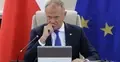 Donald Tusk z ręką przy ustach siedzi w białym fotelu. W tle flaga Polski i UE