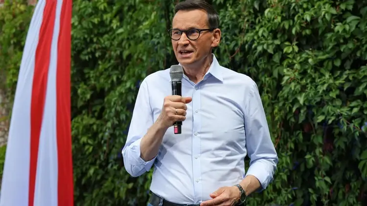 Mateusz Morawiecki przemawia do mikrofonu. W tle zielony żywopłot i polska flaga