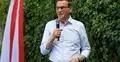 Mateusz Morawiecki przemawia do mikrofonu. W tle zielony żywopłot i polska flaga
