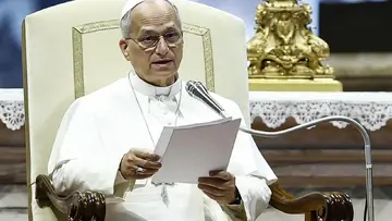 Papież Leon XIV przemawia do mikrofonu. Siedzi na fotelu, w ręku trzyma kartki