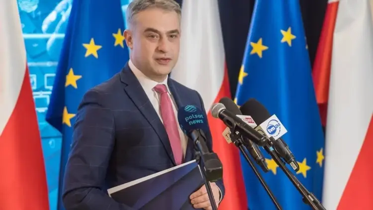 Krzysztof Gawkowski przemawia do mikrofonu na konferencji prasowej. W ręku trzyma teczkę. W tle flagi Polski i UE