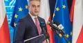Krzysztof Gawkowski przemawia do mikrofonu na konferencji prasowej. W ręku trzyma teczkę. W tle flagi Polski i UE
