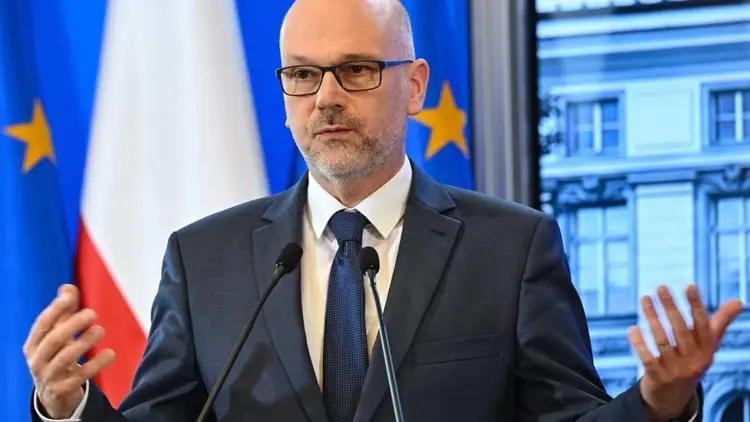 Maciej Berek przemawia i gestykuluje na konferencji prasowej. W tle flaga Polski i UE
