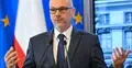 Maciej Berek przemawia i gestykuluje na konferencji prasowej. W tle flaga Polski i UE