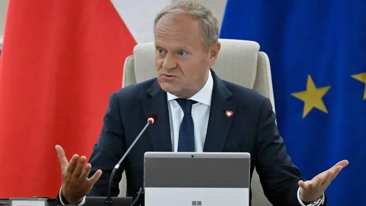 Donald Tusk gestykuluje siedząc w fotelu. W tle flaga Polski i UE