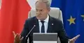 Donald Tusk gestykuluje siedząc w fotelu. W tle flaga Polski i UE