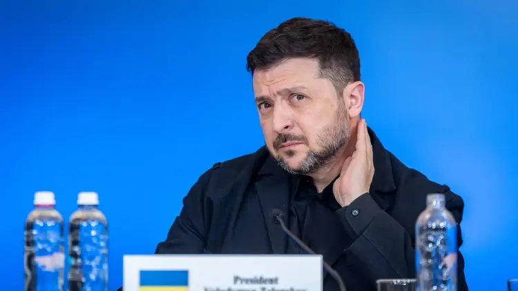 Wołodymyr Zełenski drapie się po szyi. Niebieskie tło, na stole stoi woda