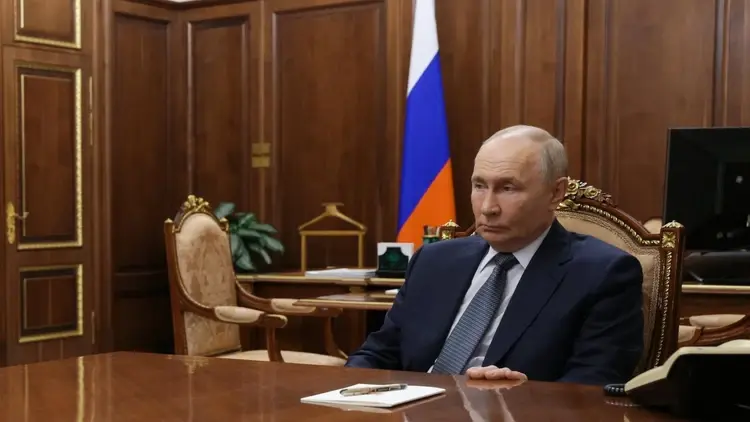 Władimir Putin siedzi przy stole z dłonią na biurku, w tle flaga Rosji