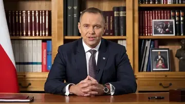 Karol Nawrocki przemawia na siedząco na tle biblioteczki i polskiej flagi