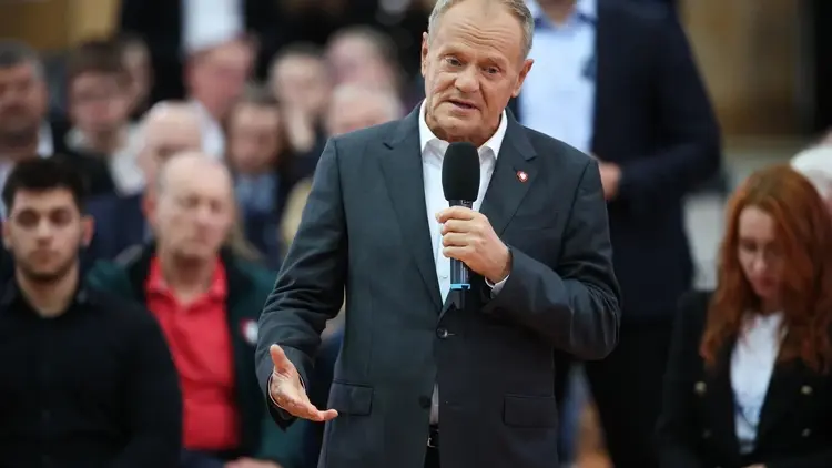 Premier Donald Tusk przemawia do mikrofonu na stojąco. W tle ludzie